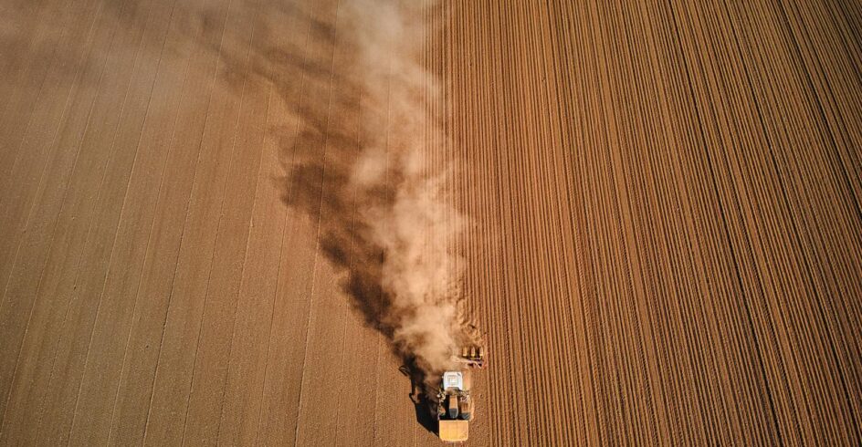 Aerial shot of a tractor ploughing a dusty field in High Peak, England, showcasing rural agriculture.